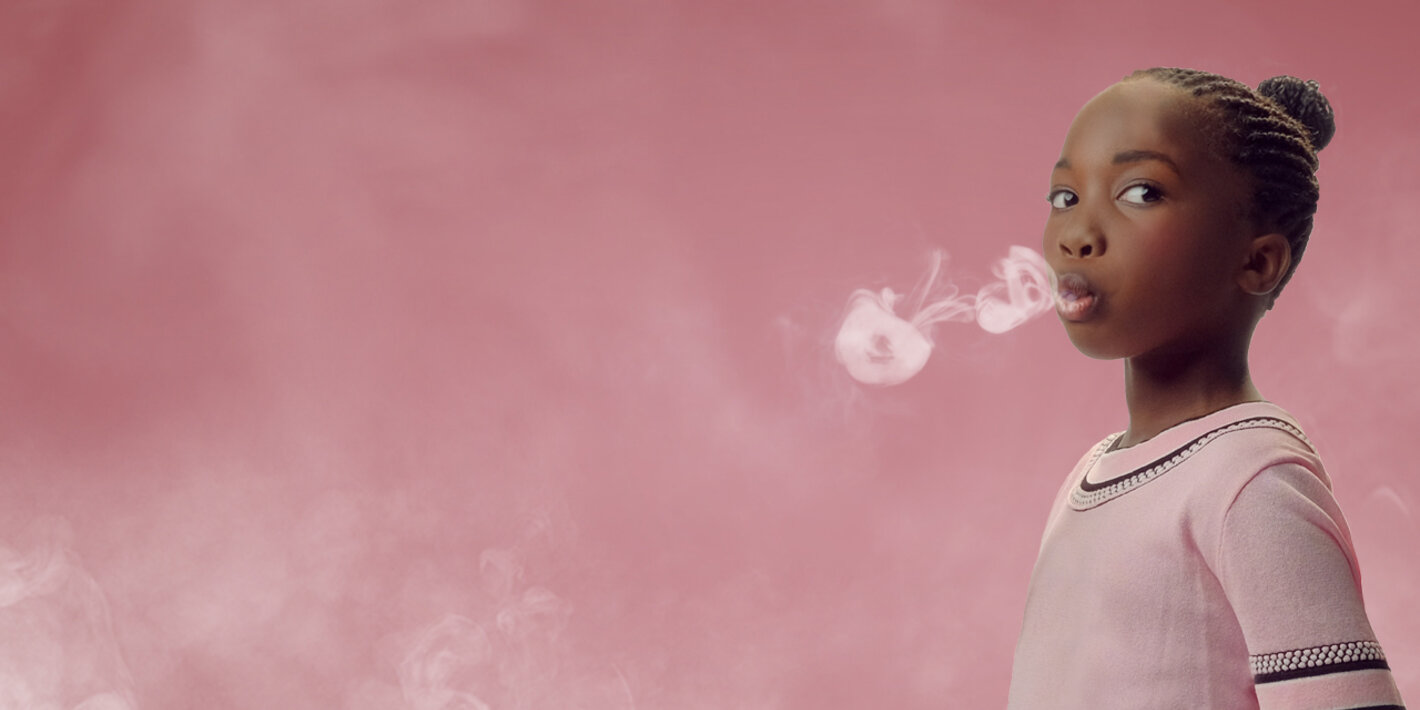 Young dark-skinned girl in pink sweater exhales smoke rings on a pink background